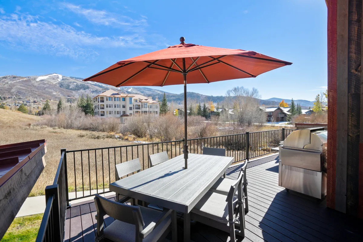 An inviting outdoor dining area featuring a table with a red umbrella, a grill, and a panoramic mountain view provides the perfect space for alfresco family meals. The scene feels tranquil and social, hinting at sunset dinners, barbecue parties, and time spent together enjoying the outdoors.