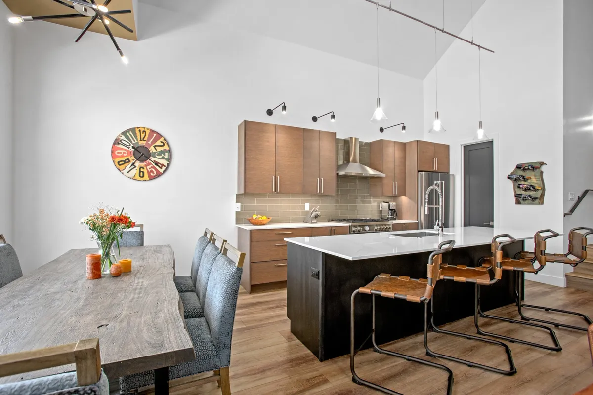 A bright, open kitchen and dining area featuring a large central island and a spacious dining table. The colorful wall clock adds a playful touch, and the arrangement encourages gatherings for both cooking and dining.