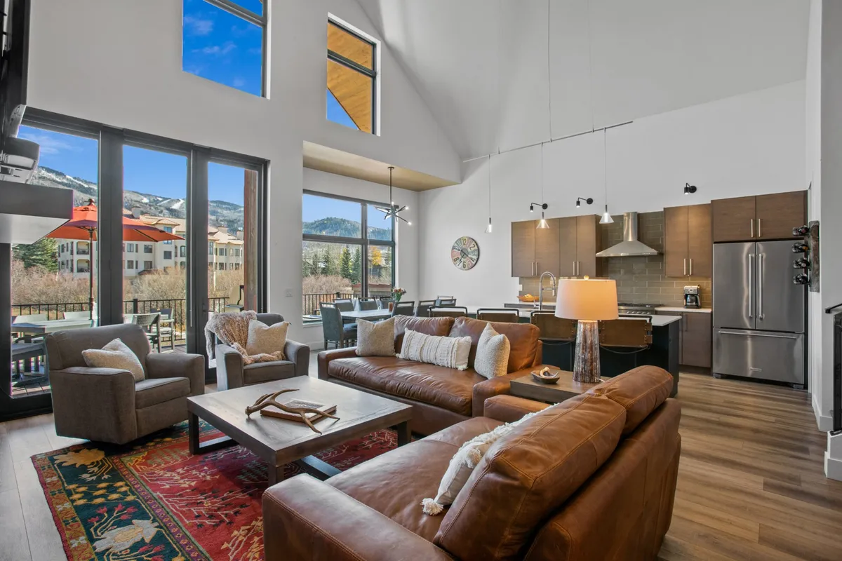 This interior shot captures an expansive open-concept living area with comfortable leather sofas, a wall of floor-to-ceiling windows showcasing sweeping mountain views, a modern dining table, and a contemporary kitchen with stainless steel appliances. The space feels airy and welcoming, emphasizing both the gathering and scenic aspects of the home.