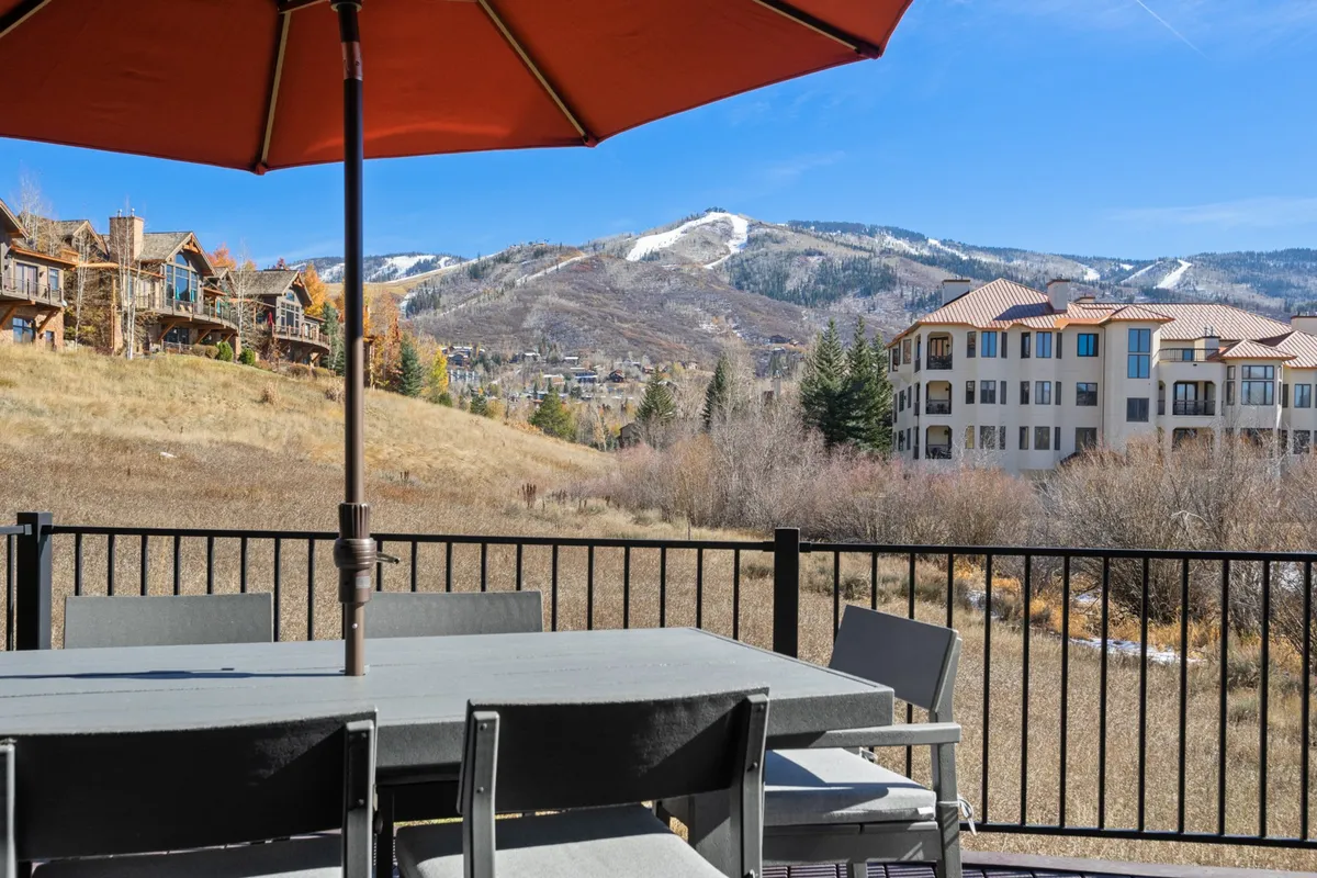 An inviting outdoor dining area with a table and umbrella set against a breathtaking mountain landscape. The setup suggests a place for group meals, drinks, or casual hangouts, with the beauty of Steamboat's outdoors all around. Other nearby buildings hint at easy access to amenities and the local scene.