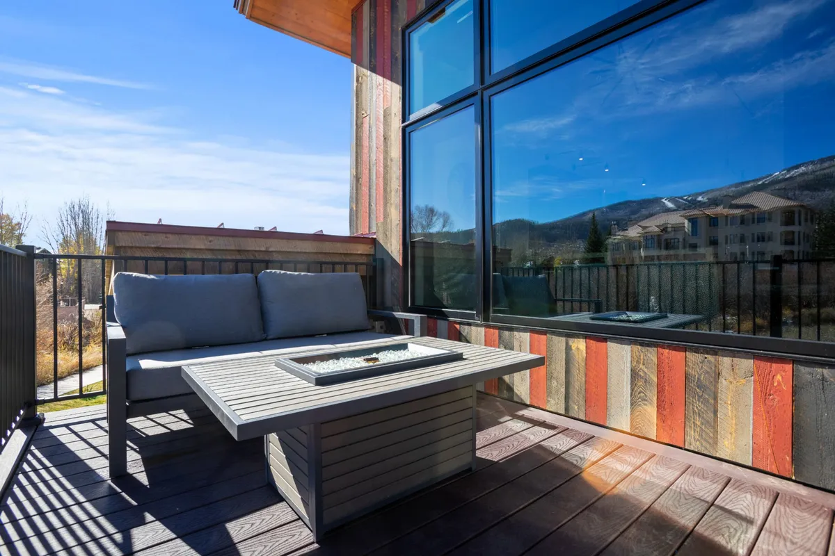 A beautiful outdoor seating area featuring a fire pit, surrounded by comfortable lounge chairs. Large windows of the residence overlook the area, and the mountain view in the background creates a stunning setting. The fire pit is the focal point, suggesting a perfect spot for après-ski gatherings and socializing in the fresh mountain air.