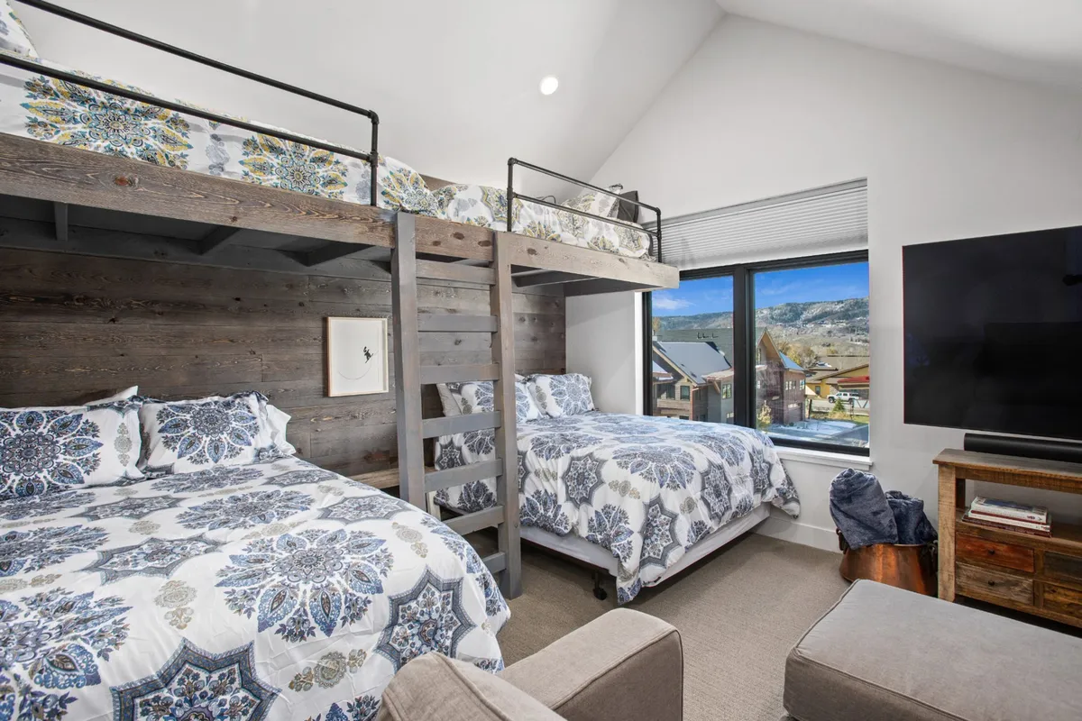 A spacious bunk room with built-in bunk beds, decorative bedding, a large window letting in natural light, and a TV. The space looks playful yet comfortable, suitable for kids, teens, or even adults.