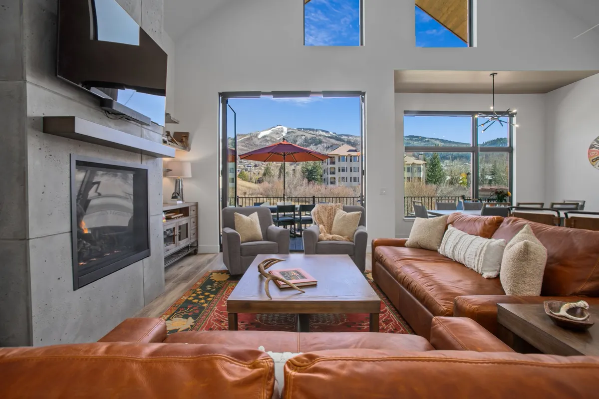 A cozy living and dining area with large windows offering views of the mountains, plush leather sofas, a modern fireplace, and an elegant dining table. The ambiance is inviting, with the fireplace providing a focal point for relaxation and socializing.