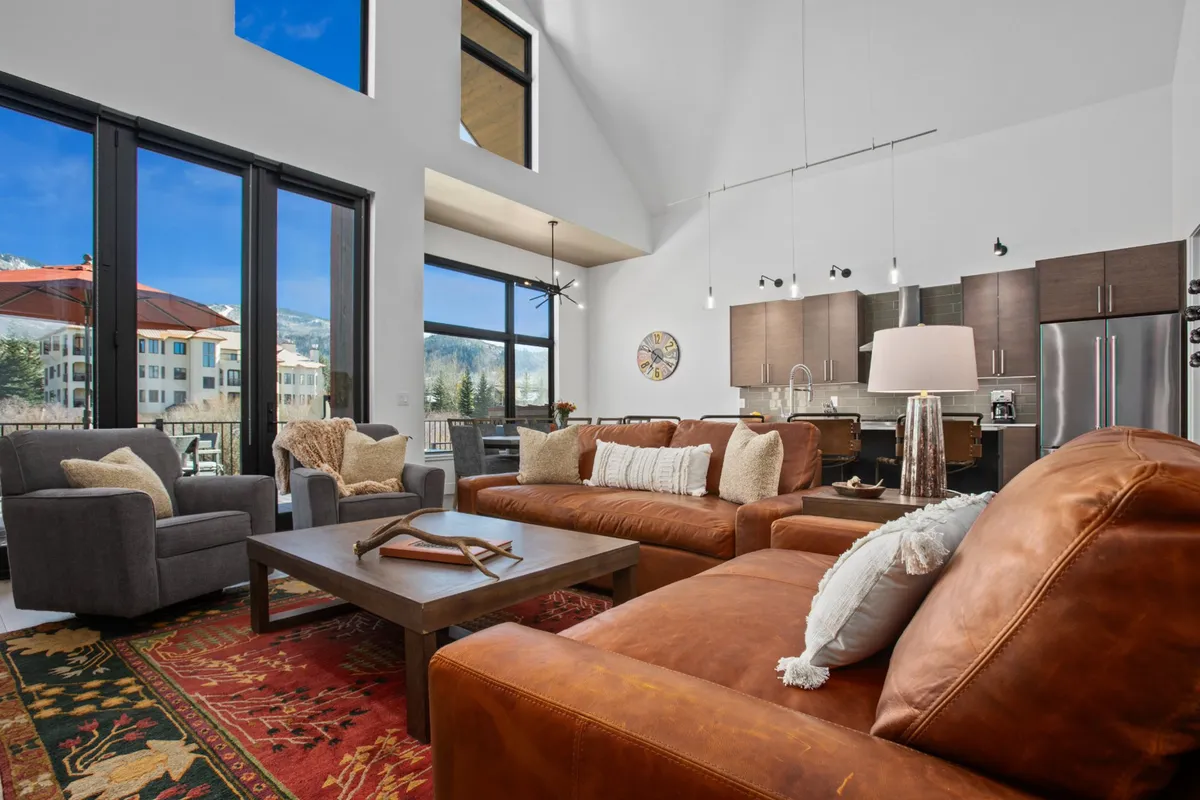 A spacious and welcoming interior living area with large windows framing mountain views, leather sofas arranged for conversation, a coffee table with decorative accents, and a modern kitchen in the background. The space is designed for socializing, movie nights, and relaxing together after a day out.