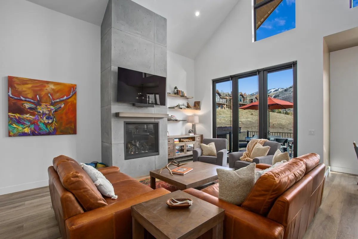 A modern living area with plush leather sofas and a fireplace is accentuated by large windows showcasing the mountain landscape. Artwork on the walls and the warm glow of the fire create a cozy yet luxurious ambiance.