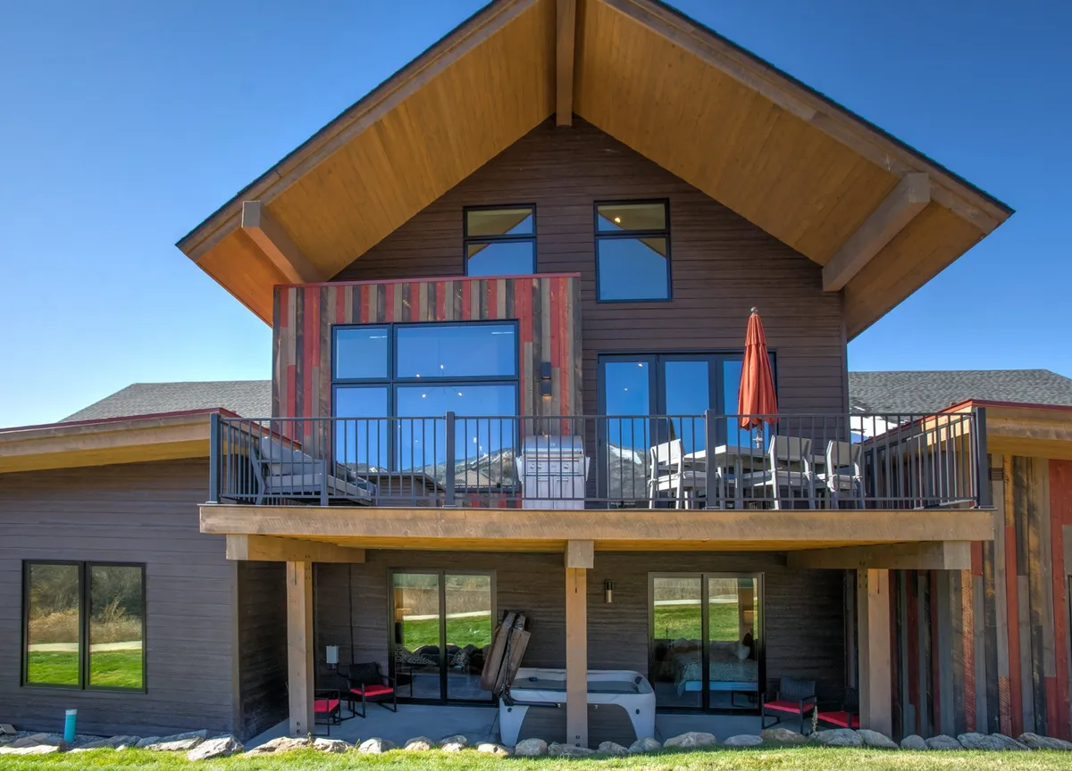 The image shows the exterior of a newly constructed mountain modern home in Steamboat Springs. Large windows allow natural light to flood the interior, while a spacious wooden deck is furnished with inviting outdoor seating. The colorful siding adds vibrancy, and the deck overlooks scenic mountain surroundings, perfect for relaxing or entertaining guests. The setting suggests moments of stargazing or sipping morning coffee amidst the fresh mountain air.