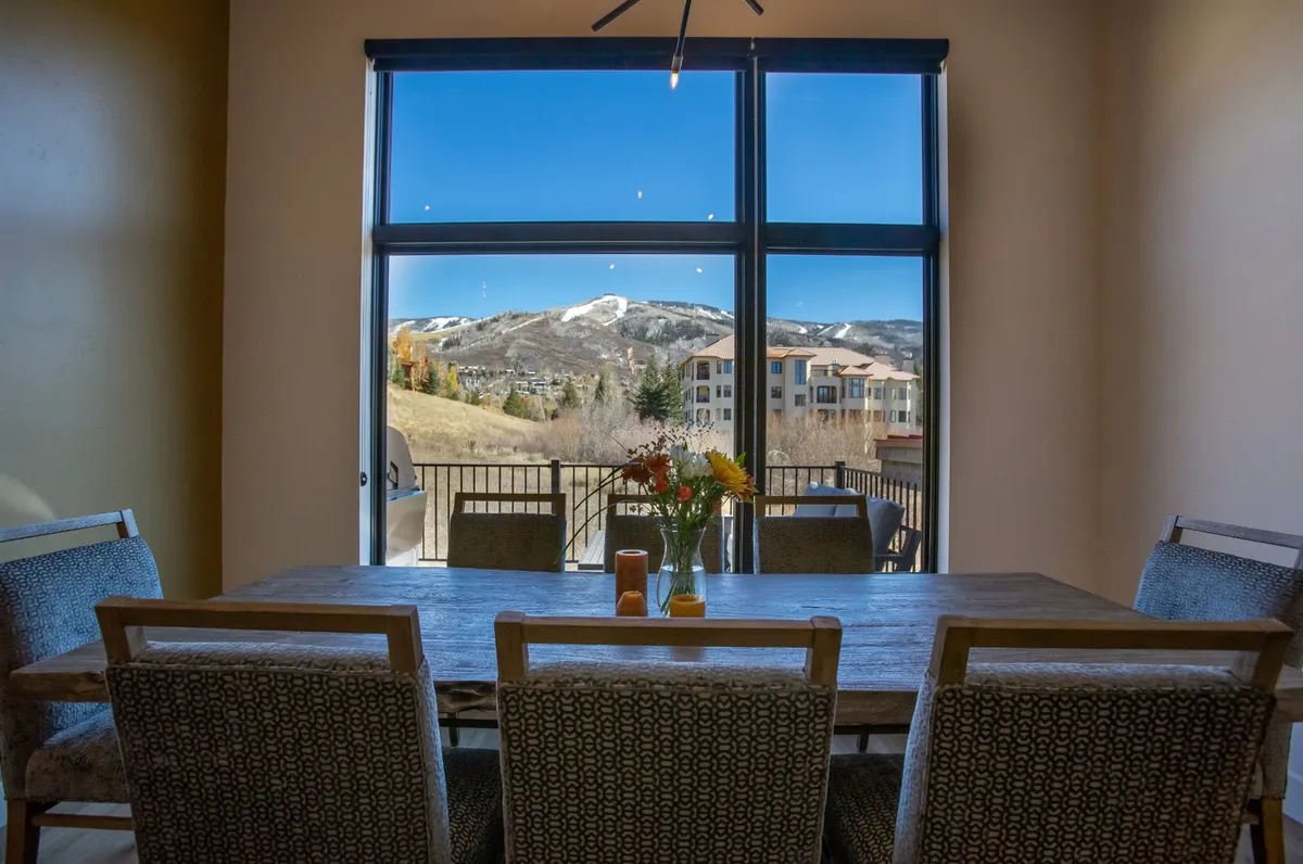 A bright dining area with a large table sits beside expansive windows that frame a stunning mountain vista. The space is filled with natural light, and a flower vase adds a touch of elegance to the table, inviting guests to sit and enjoy the view.
