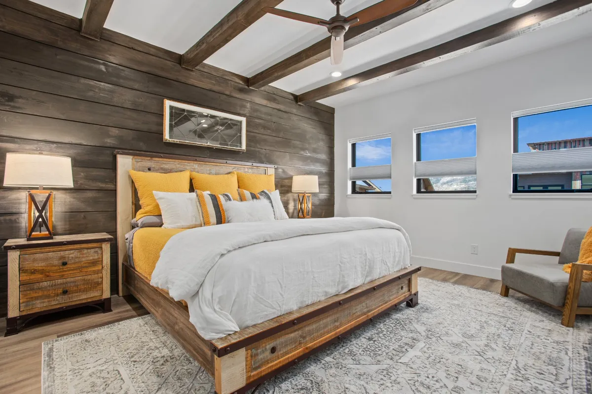 A warmly lit king bedroom featuring a comfortable bed with yellow accent pillows, a nightstand, and a soft rug. Natural light streams in, creating a welcoming and restful atmosphere ideal for relaxation.