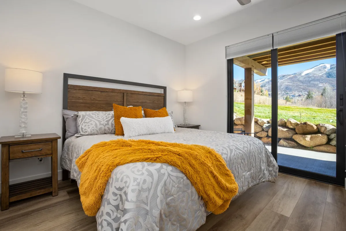 A cozy bedroom features a bed with decorative pillows and a large glass door that opens to the outside, providing easy access to fresh air and scenic views. A nightstand with a lamp sits nearby, completing the inviting atmosphere.