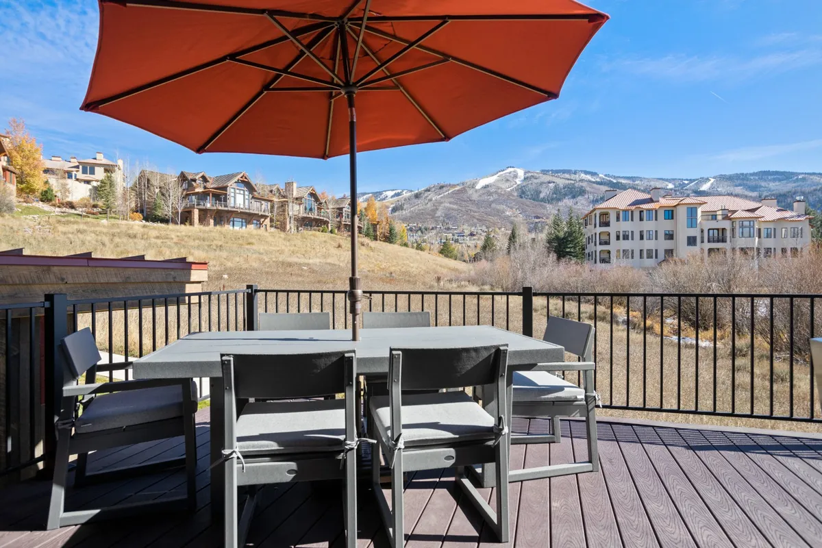 A spacious outdoor deck features a dining table under a vibrant orange umbrella, set against a backdrop of stunning mountain vistas. The space is designed for al fresco dining, relaxation, and social gatherings, embodying the home's connection to the outdoors.