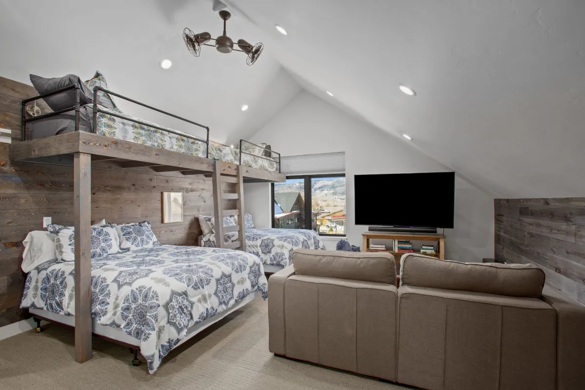 A cozy, spacious bunk room with multiple bunk beds, large windows, a sofa, and a television. The setup is designed for children or multiple family members to gather, play games, watch TV, or share stories, with sunlight streaming in through the windows.